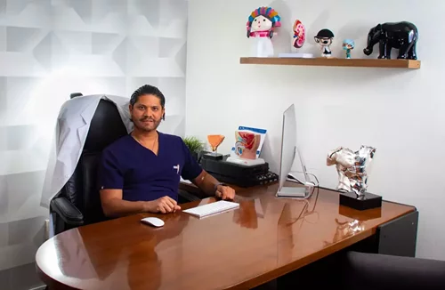 urologo_consultorio_rene_zamora_queretaro_326x500 Dr Rene Zamora sentado en su consultorio urologico en Queretaro, junto a simbolos decorativos y elementos institucionales.